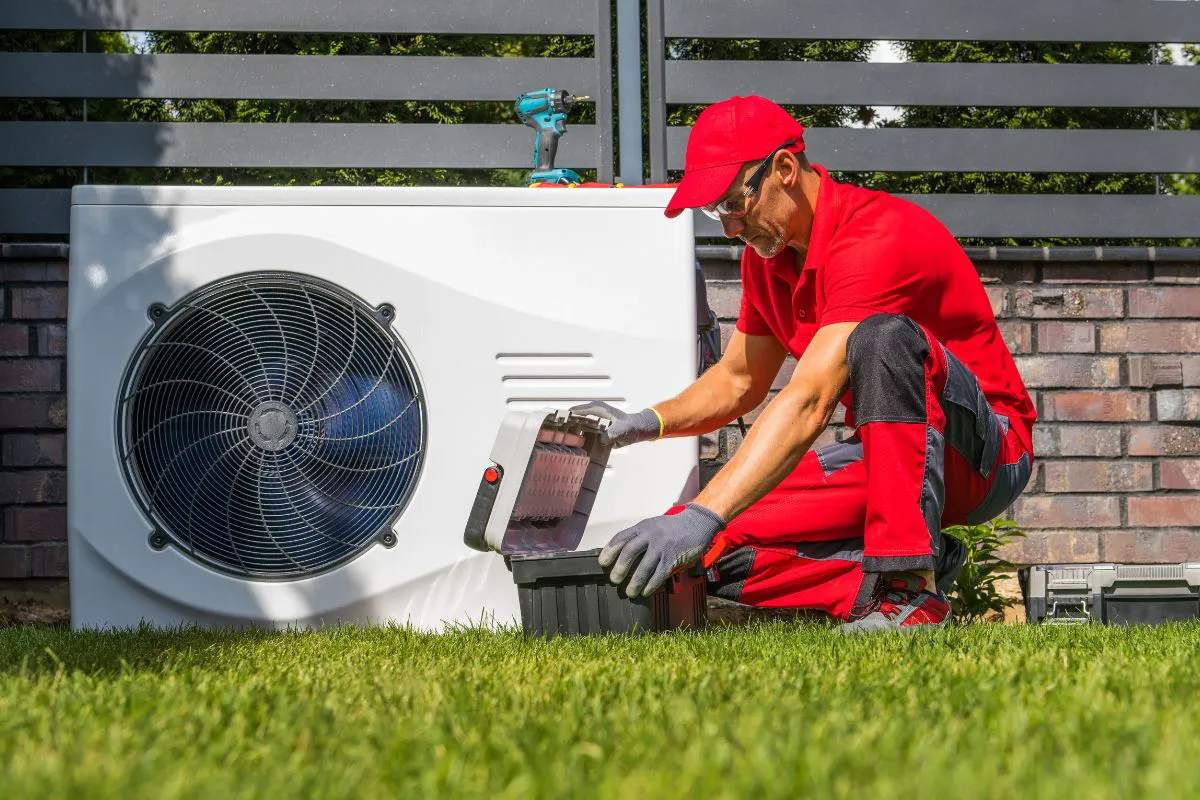 Warmtepompinstallatie buiten wordt geïnspecteerd door monteur in rode werkkleding bij nieuwbouwwoning.